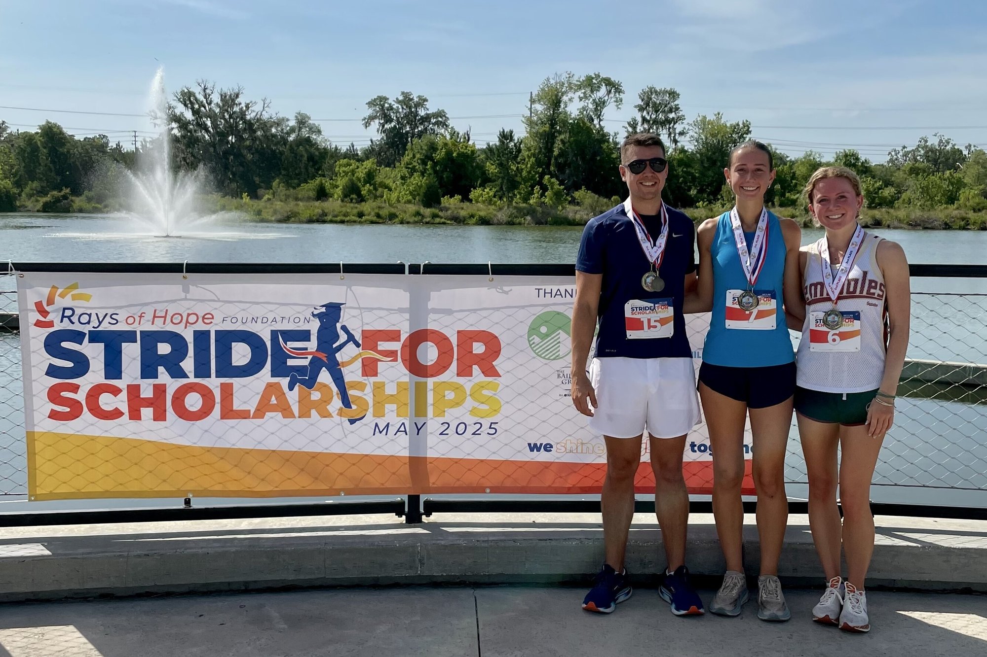 Runners posing with medals beside the Rays of Hope Stride for Scholarships 5K banner at the charity fun run.