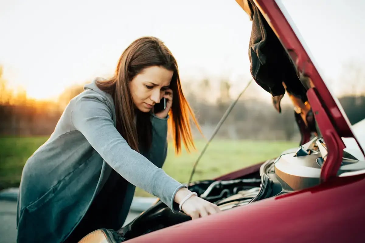 Lady on the phone while looking under the hood of a parked car.