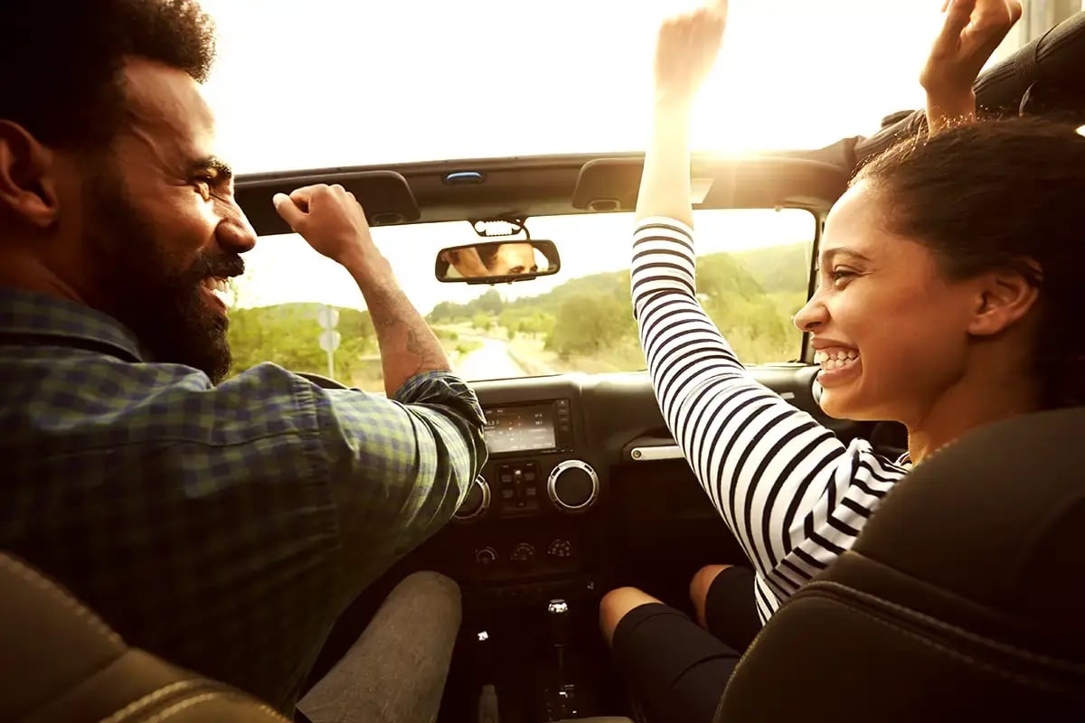 Couple rides in convertible raising arms excitedly in the air with sunlight